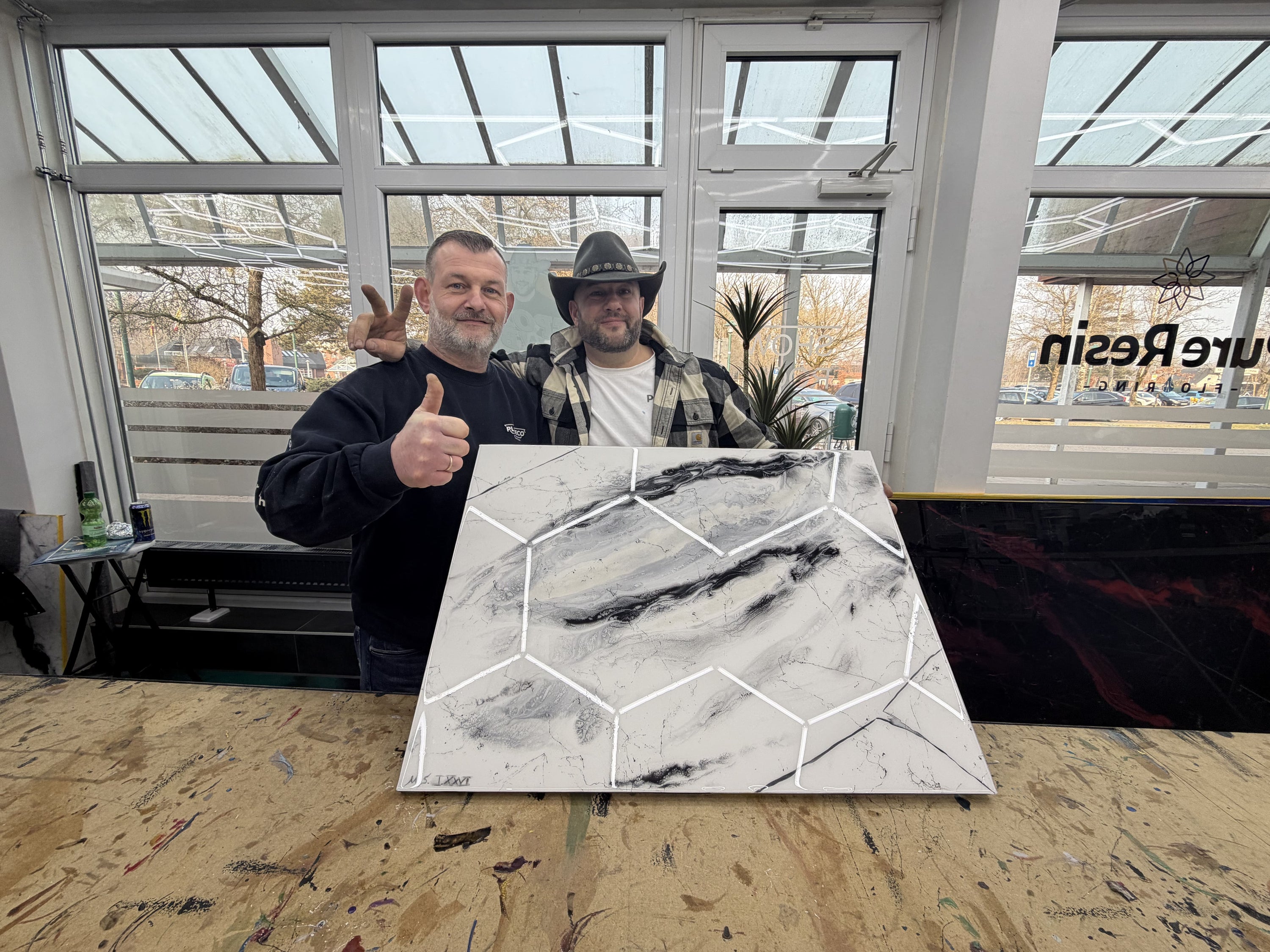 Teilnehmer eines Epoxidharz-Workshops präsentieren stolz eine selbst gestaltete Musterplatte in weißer Marmoroptik mit schwarzen Adern.
