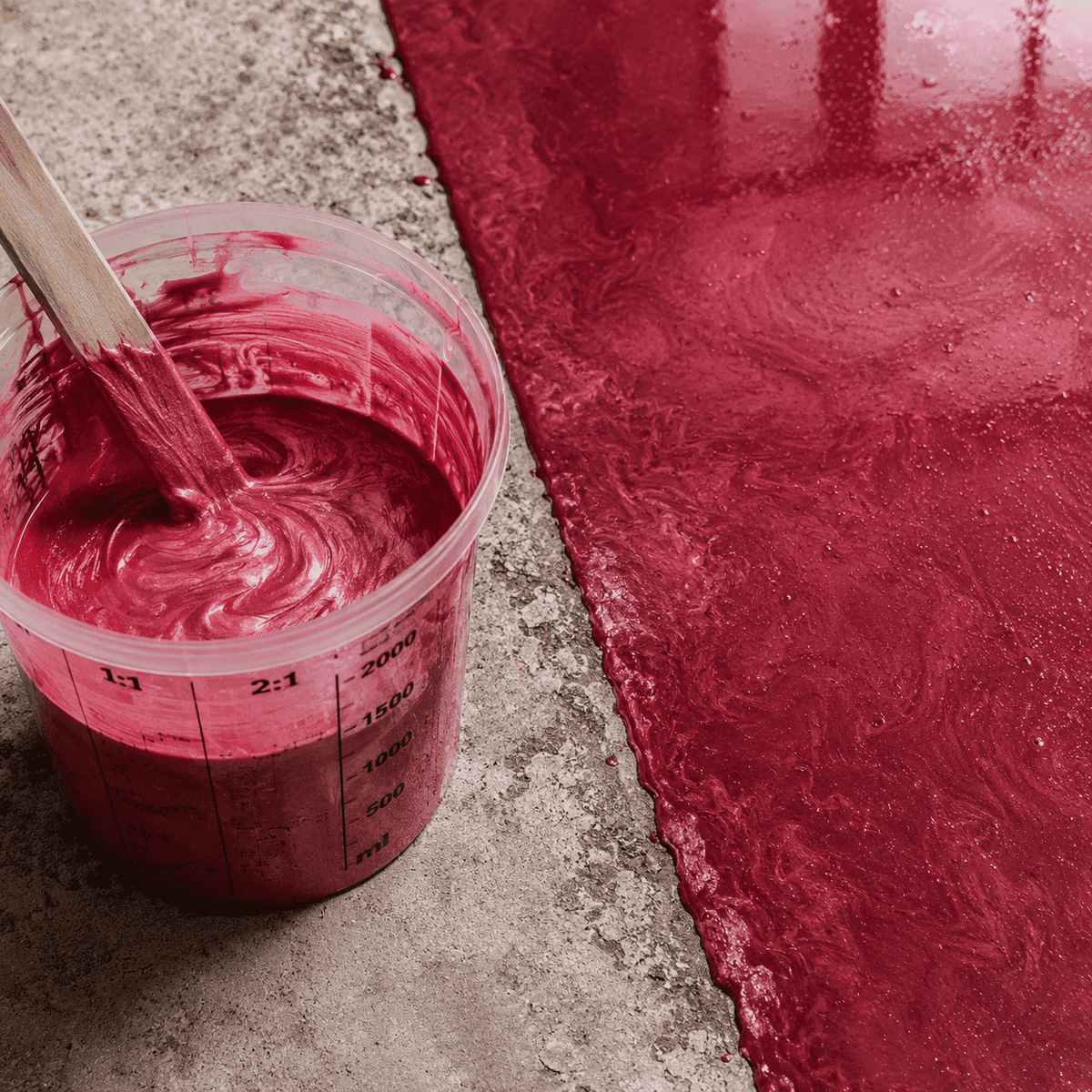 Mauve-rotes Metallic-Epoxidharz im Mischbecher neben einer frisch beschichteten Betonfläche mit intensivem Perleffekt, edler Marmorstruktur und glänzender Tiefenwirkung.