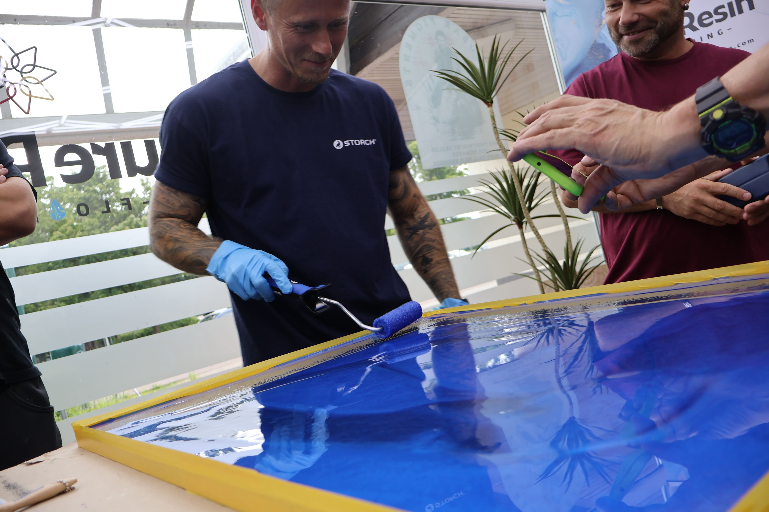 Teilnehmer eines Epoxidharz-Workshops versiegelt eine blaue Metallic-Epoxidharz-Musterplatte mit einer Rolle unter Anleitung des Trainers.