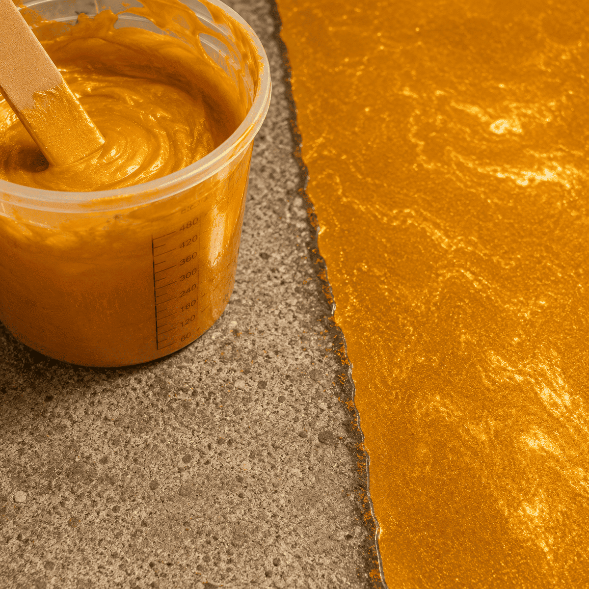 Olympic-Gold Metallic-Epoxidharz im Mischbecher neben einer frisch beschichteten Betonfläche mit intensivem Schimmer- und Marmoreffekt.
